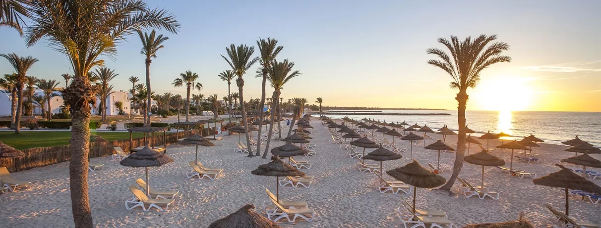 Plage de Djerba au crépuscule avec parasols en paille et transats face à la mer Méditerranée.