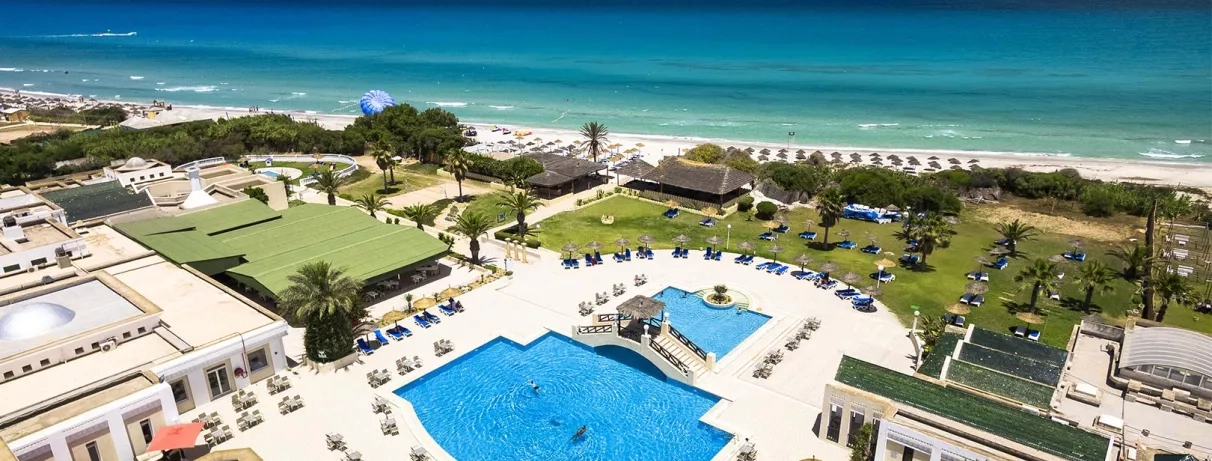 Vue aérienne de la piscine principale de l’hôtel, entourée de jardins verdoyants, avec la plage et la mer turquoise en arrière‑plan