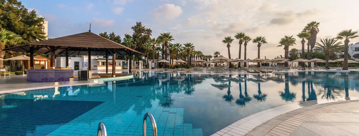 Grande piscine extérieure de l'hôtel Marhaba Palace bordée de palmiers et parasols sous un ciel azur à Monastir.
