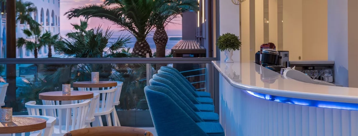 bar, fauteuils de bar bleus, petites tables rondes en bois avec chaises blanches, baie vitrée ouverte sur l'extérieur, vue sur la mer et les palmiers