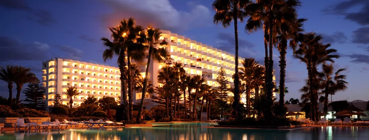 Façade illuminée de l'hôtel Sahara Beach Monastir et piscine de nuit pour un séjour balnéaire.