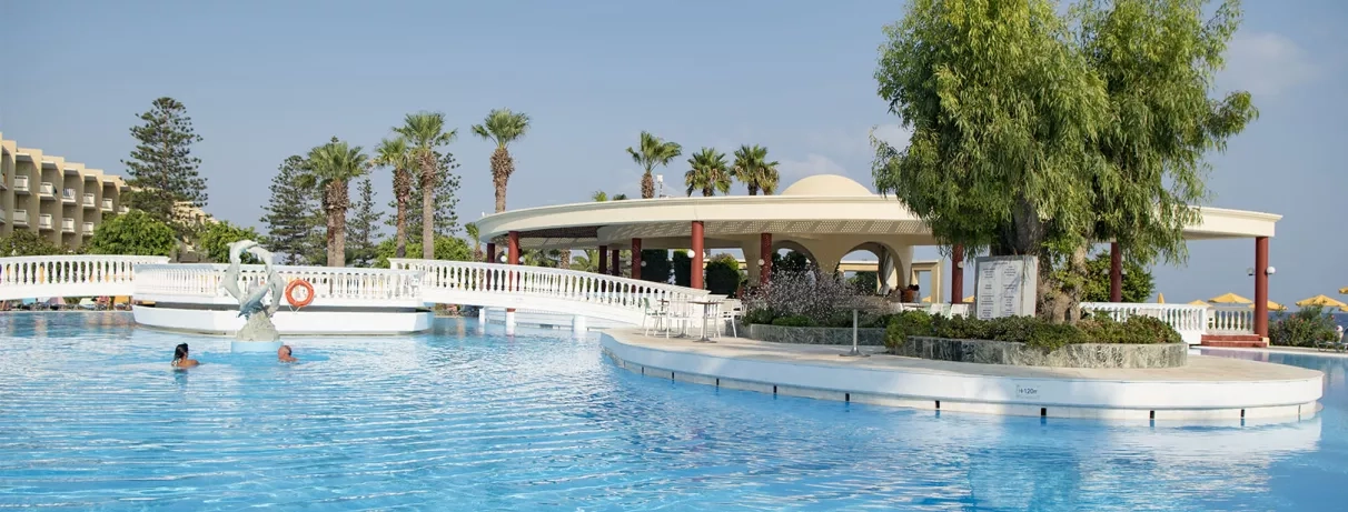 piscine pour adultes traversée par un pont permettant d'accéder au bar terrasse et à la plage, personnes qui se baignent, sculpture de dauphins, arbres et palmiers 