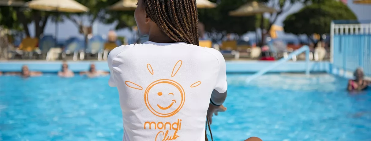 animatrice de dos avec tee shirt mondi club, assise devant la piscine de l'hôtel