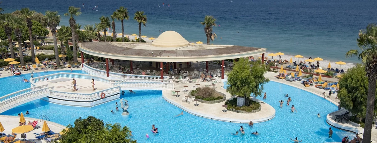 vue aérienne mer et piscine, bar de la piscine, pont et plateforme permettant de traverser la piscine, accès plage de galets, palmiers, transats et parasols