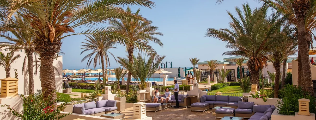 Salon extérieur confortable avec canapés entouré de palmiers dans les jardins luxueux de l'hôtel Ulysse Djerba.