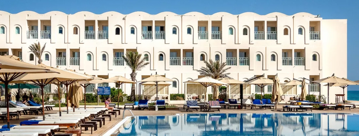 Façade blanche de l'hôtel Ulysse Djerba se reflétant dans l'eau cristalline de la piscine sous le soleil.