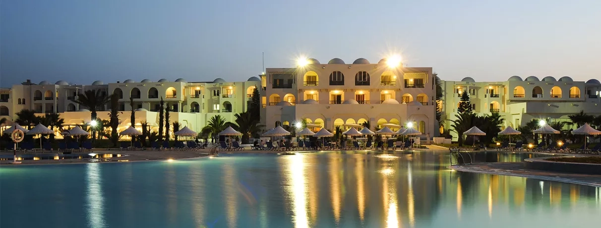 Vue nocturne illuminée de la piscine et des façades blanches de l'hôtel en bord de mer.