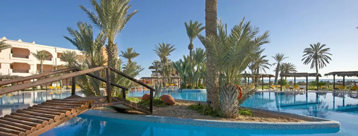 Grande piscine lagon bordée de palmiers et pont en bois sous le soleil de l'hôtel Safira Palms Djerba.
