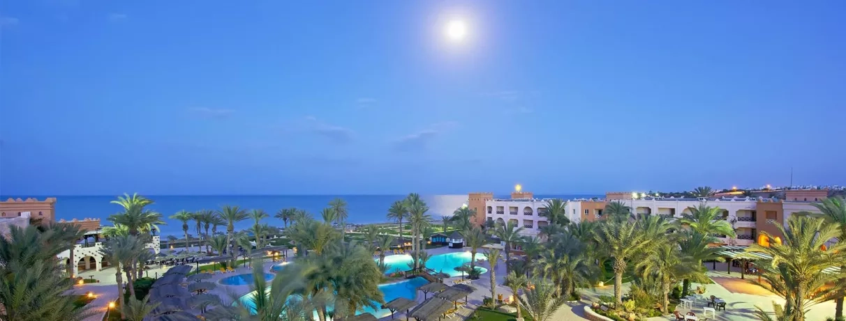 Paysage nocturne du resort Safira Palms Djerba avec piscines éclairées, palmiers et vue sur mer sous la lune.