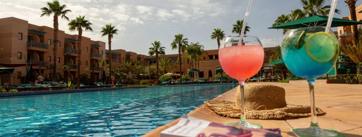 piscine, deux cocktails orange et bleu posés sur le rebord de la piscine, palmiers, hôtel en fond, livre et chapeau de paille