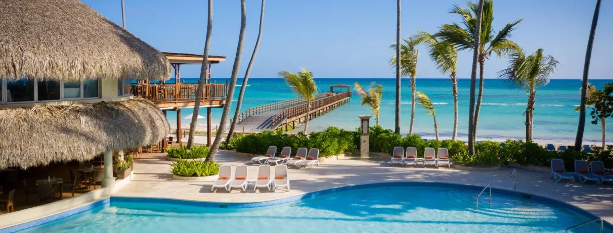 Plage avec ponton, sable, eau claire, transats blancs autour de la piscine, palmiers, pieds dans l'eau