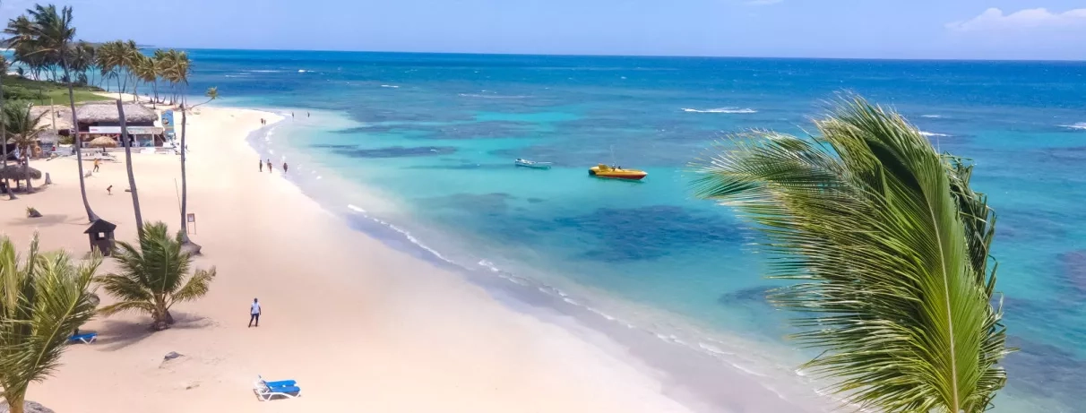 Vue plage de sable blanc, palmiers, eaux turquoises, transats bleus, bateaux au large