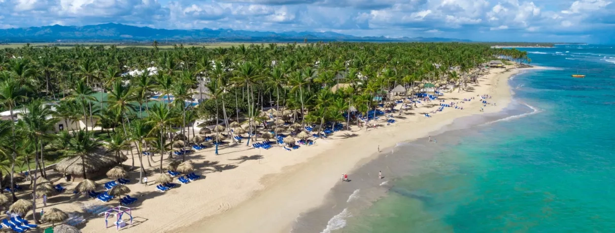 Vue plage de sable blanc, eau turquoise, palmiers, transats bleus, piscine