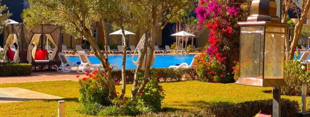 en premier plan, le jardin de l'hôtel avec des arbres et en deuxième plan,  la piscine et les transats