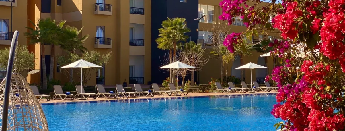 piscine en premier plan avec les transats juste derrière et au fond le bâtiment des chambres