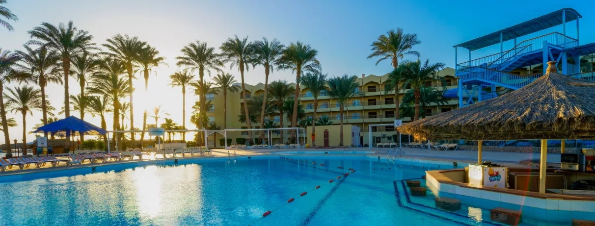 vue piscine extérieur avec bar central, transats autour, coucher de soleil, palmier, bâtiment de l'hôtel