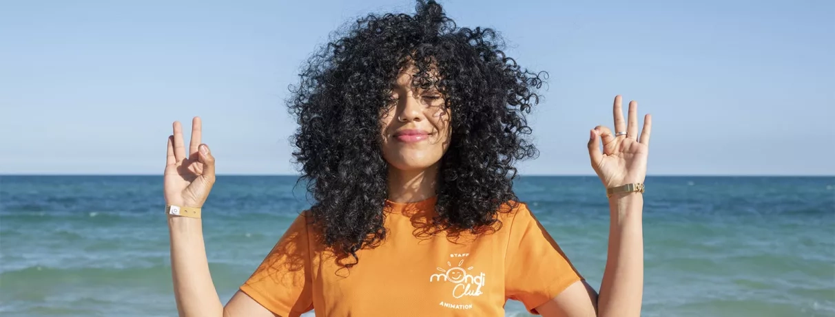 animatrice brune mondi club qui ferme les yeux, tient ses mains en position de yoga sur la plage devant la mer, ambiance zen