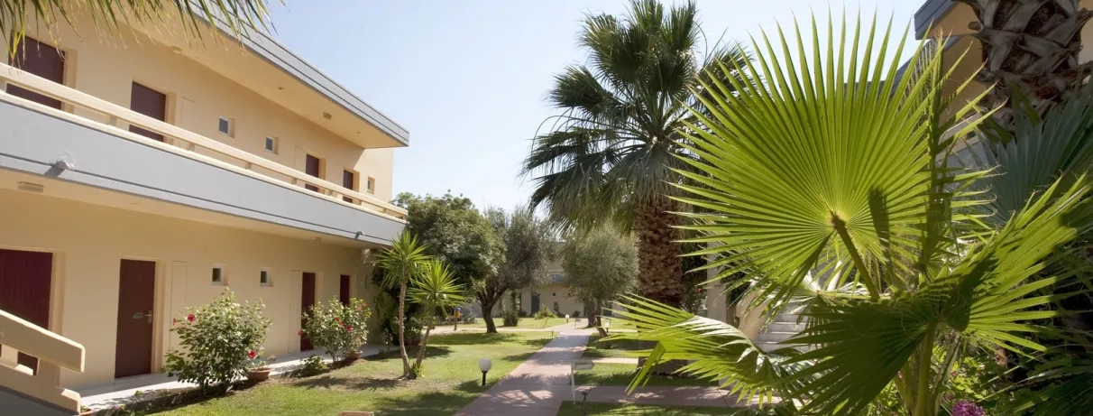 allée avec palmiers, végétation luxuriante, lampe, bâtiments de l'hôtel avec un étage et escalier, ciel bleu