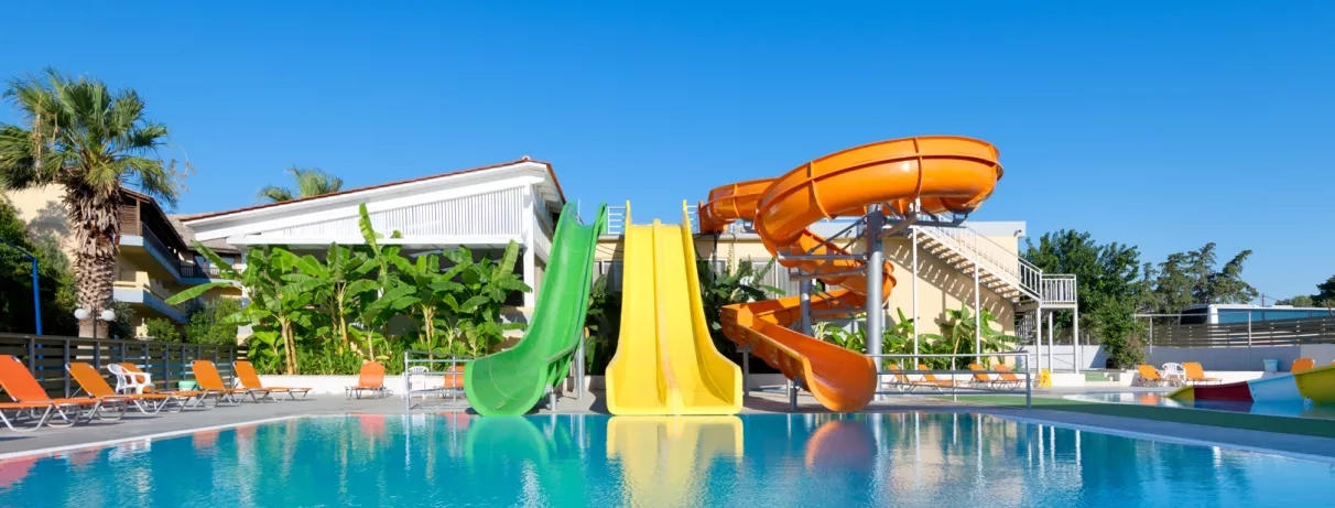aquapark, 3 toboggans vert jaune orange, piscine, transats oranges, végétation, palmiers, ciel bleu