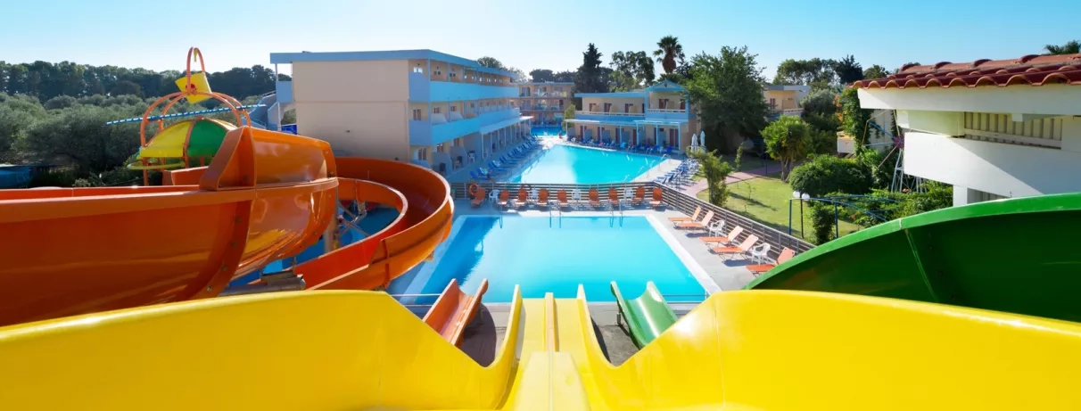 toboggans, deux piscines et transats autour, végétation, plusieurs bâtiments avec deux étages, ciel bleu