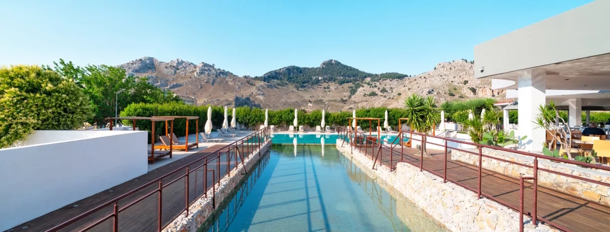 piscine couloir, lits structure en bois, parasols et transats blancs, restaurant terrasse sur la droite et montagne en arrière-plan