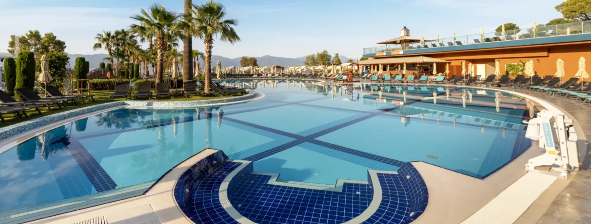 Une vaste piscine ensoleillée, bordée de palmiers et de transats sous un ciel bleu.