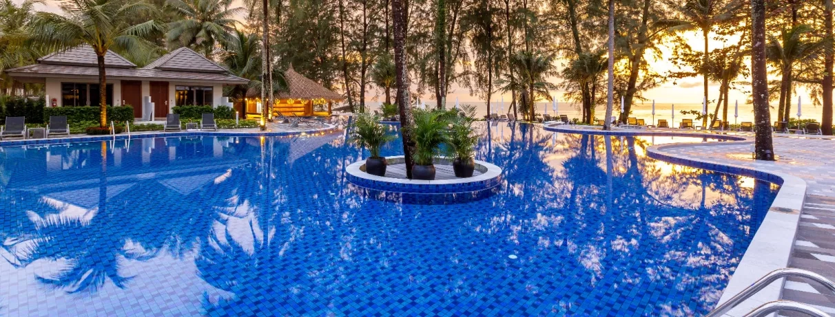 Une large piscine entourée de palmiers et de jardins tropicaux au coucher du soleil.
