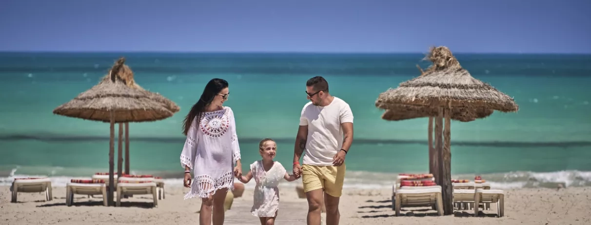 Famille marchant sur une plage de sable fin bordée de parasols typiques aux Quatre Saisons à Djerba.
