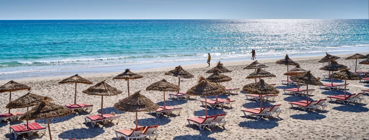 Plage privée avec rangées de parasols en paille et chaises longues face à la mer Méditerranée, Djerba.