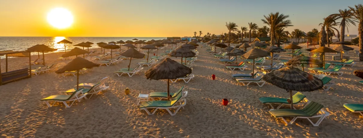Plage de sable fin au coucher du soleil avec parasols en paille et transats à Djerba, Tunisie.