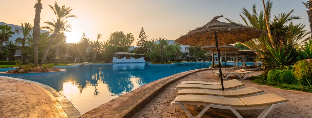 Grande piscine extérieure au coucher du soleil avec transats et parasols de paille au Djerba Resort.