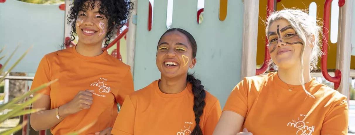 Trois animatrices souriantes portant des t-shirts orange et du maquillage festif.