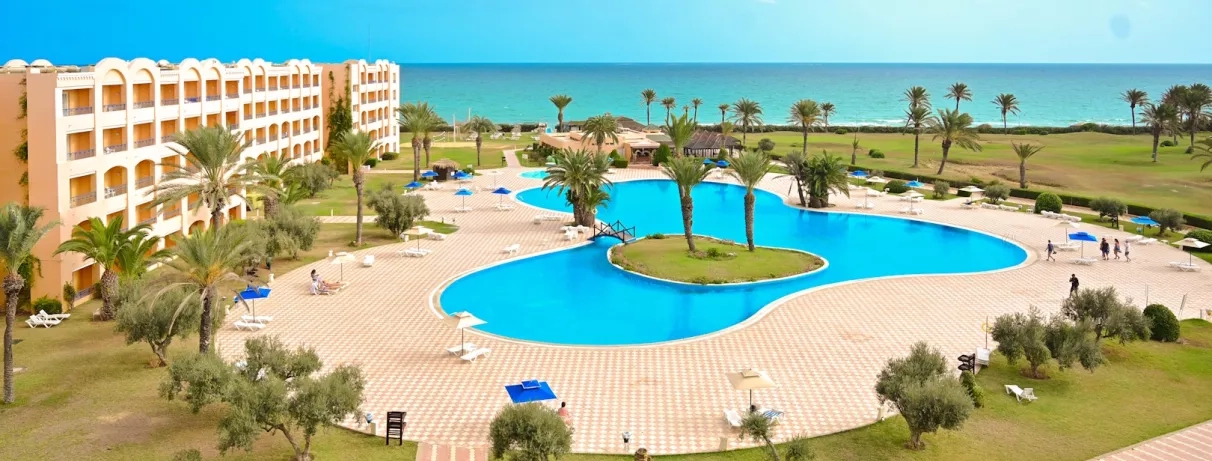 Vue globale sur la piscine extérieure principale de l’hôtel, avec un bâtiment situé sur la gauche et la mer en arrière‑plan.