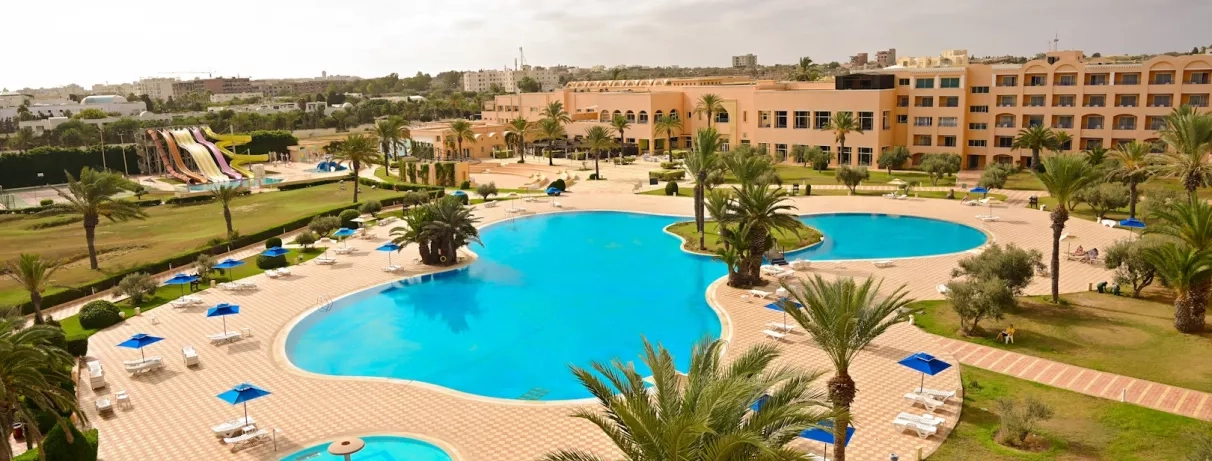 Vue d’ensemble sur la grande piscine extérieure, avec le bâtiment de l’hôtel et les toboggans aquatiques visibles en arrière‑plan