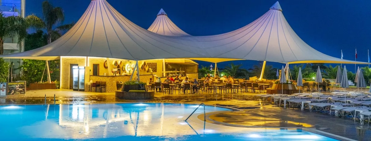 Un restaurant en bord de piscine illuminé de nuit sous de grandes tentes blanches élégantes.