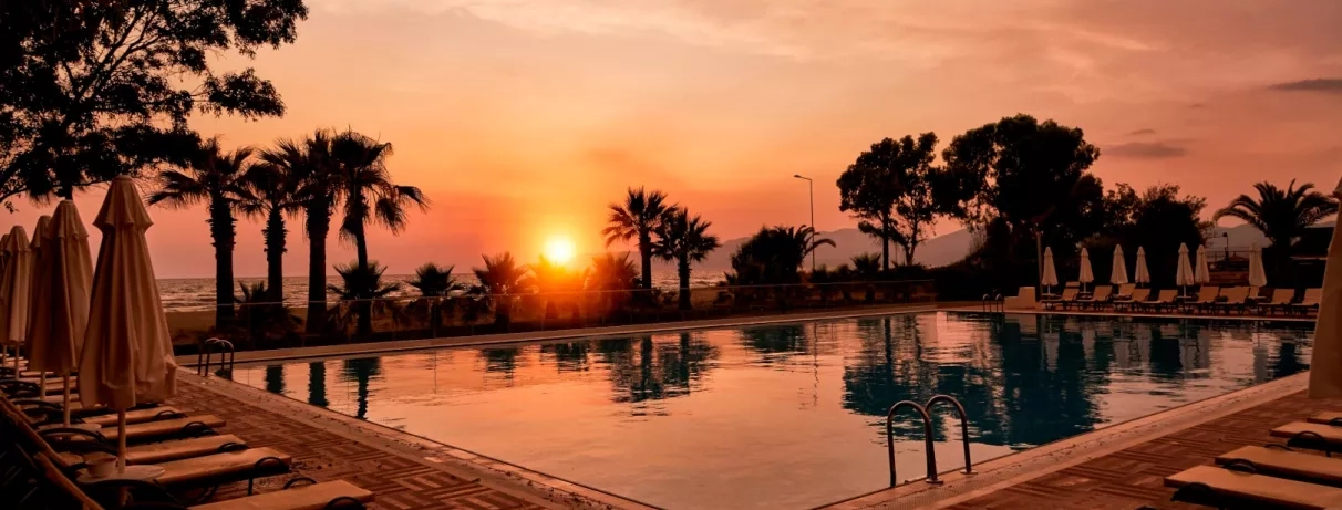 Une vue du coucher du soleil, où les palmiers et les parasols se reflètent dans l'eau de la piscine.