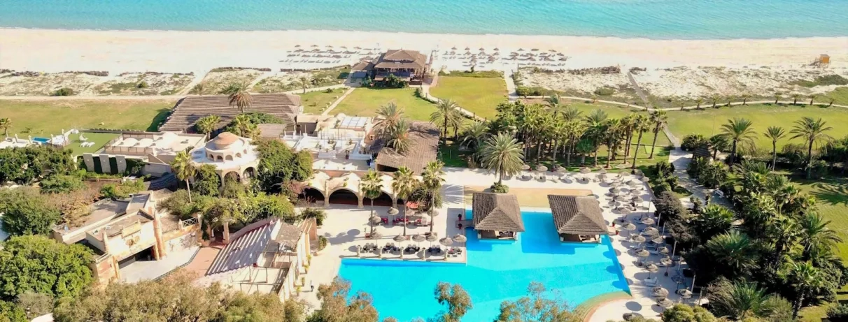 Vue aérienne d’un resort en bord de mer avec grande piscine, plage aménagée et palmiers.