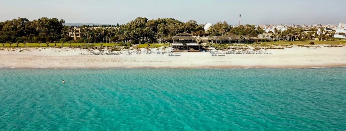 Plage de sable avec eau turquoise, transats et palmiers devant un resort entouré de verdure. 