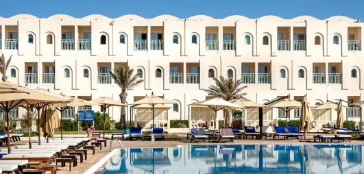 Façade blanche de l'hôtel Ulysse Djerba se reflétant dans l'eau cristalline de la piscine sous le soleil.