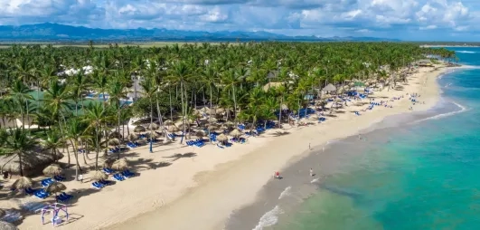 Vue plage de sable blanc, eau turquoise, palmiers, transats bleus, piscine