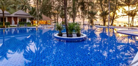 Une large piscine entourée de palmiers et de jardins tropicaux au coucher du soleil.
