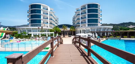 Un ponton en bois mène vers deux bâtiments d'hôtel modernes entourés de grandes piscines bleues.