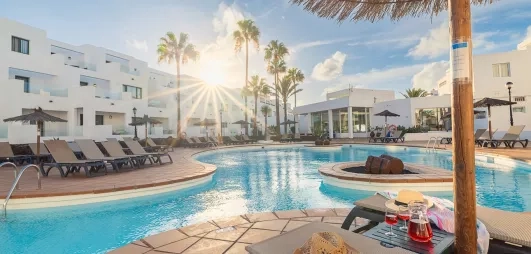 Une vue panoramique d'une grande piscine, bordée de transats et de bâtiments blancs modernes.