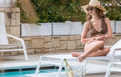 femme avec chapeau au bord de la piscine, transats blancs, bouteille de champagne, pastèque et autres fruits