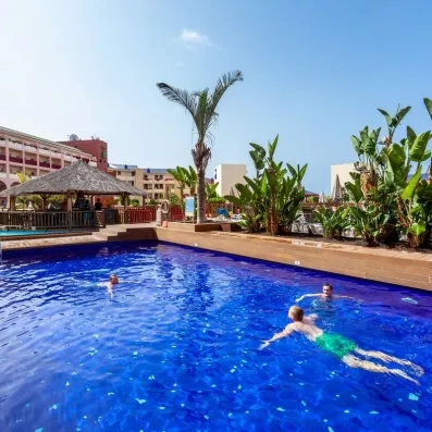 Une piscine extérieure ensoleillée, entourée de palmiers et de vacanciers.