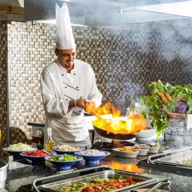 Un cuisinier prépare un plat en réalisant une cuisson flambée dans une poêle, laissant s’élever une flamme spectaculaire