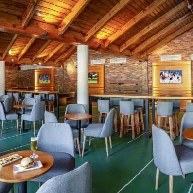 salle de restaurant chaises grises et tables rondes en bois, bière et burgers posés sur une table, toit en bois poutres apparentes, écrans diffusant du sport au mur
