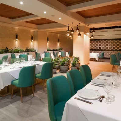 salle de restaurant chaises vertes longues tables nappes blanches, pilier et luminaires au centre, beaucoup de plantes 
