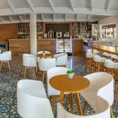 bar avec tables rondes en bois et chaises blanches, chaises hautes sur la droite, bar au fond pour petit déjeuner et gouter avec boissons chaudes, mais aussi bouteilles alcool