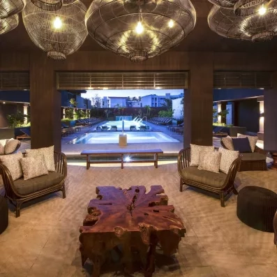 lobby avec vue piscines et jets d'eau, fauteuils confortables, table basse en bois sculpté, banc en bois, gros luminaires ronds 